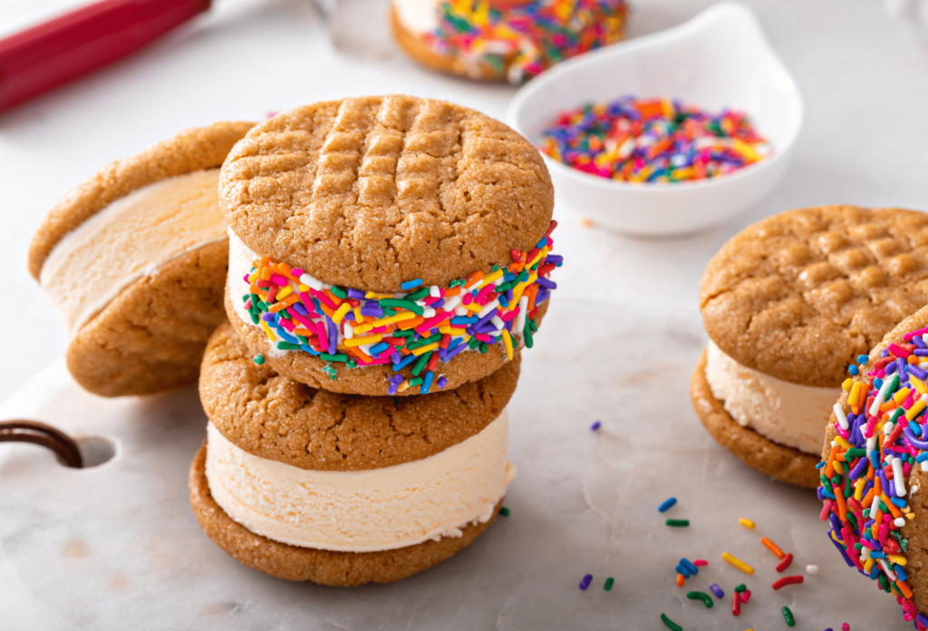 The image shows 5 ice cream sandwiches made from peanut butter cookies and vanilla ice cream. two of the sandwiches are decorated with rainbow sprinkles.