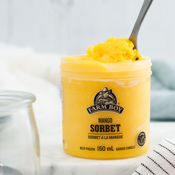 The image shows a single serve container of Farm Boy Mango Sorbet with the lid off and a spoon scooping some of it out. In the lower right corner is the white dishcloth striped with rows of tiny black squares. In the lower left corner is a glass container.