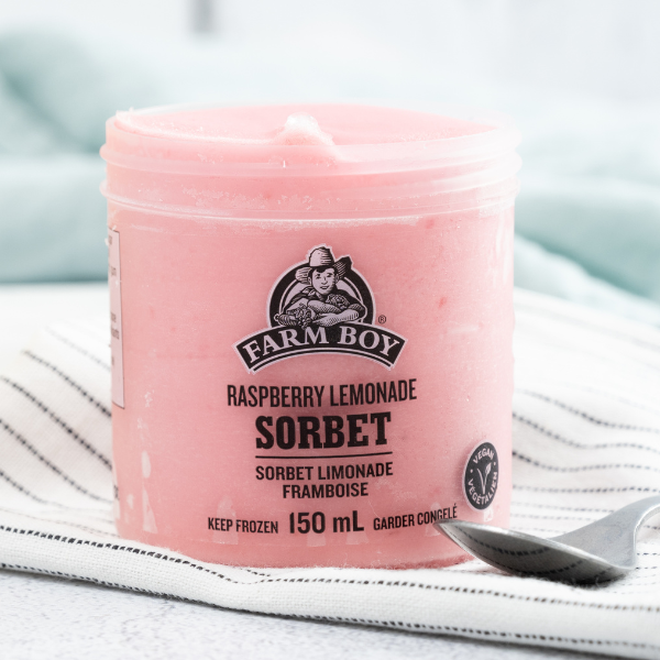 The image shows a single serve container of Farm Boy Raspberry Lemonade Sorbet with the lid off. The container is resting on a white dish cloth striped with rows of tiny black dots. There is a spoon beside the container.