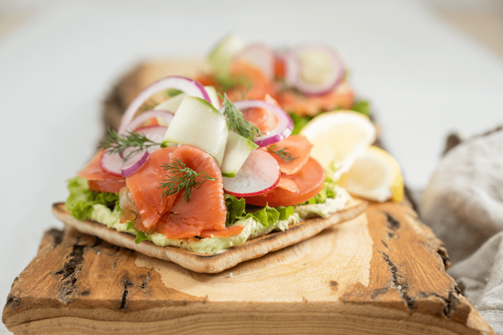 Theimage shows an open-faced sockeye salmon sandwich on a wooden cutting board. The sandwich is layered with avocado, salmon, and thinly sliced onions.