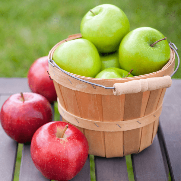 Wooden bushel of Granny Smith apples with three red apples beside it.