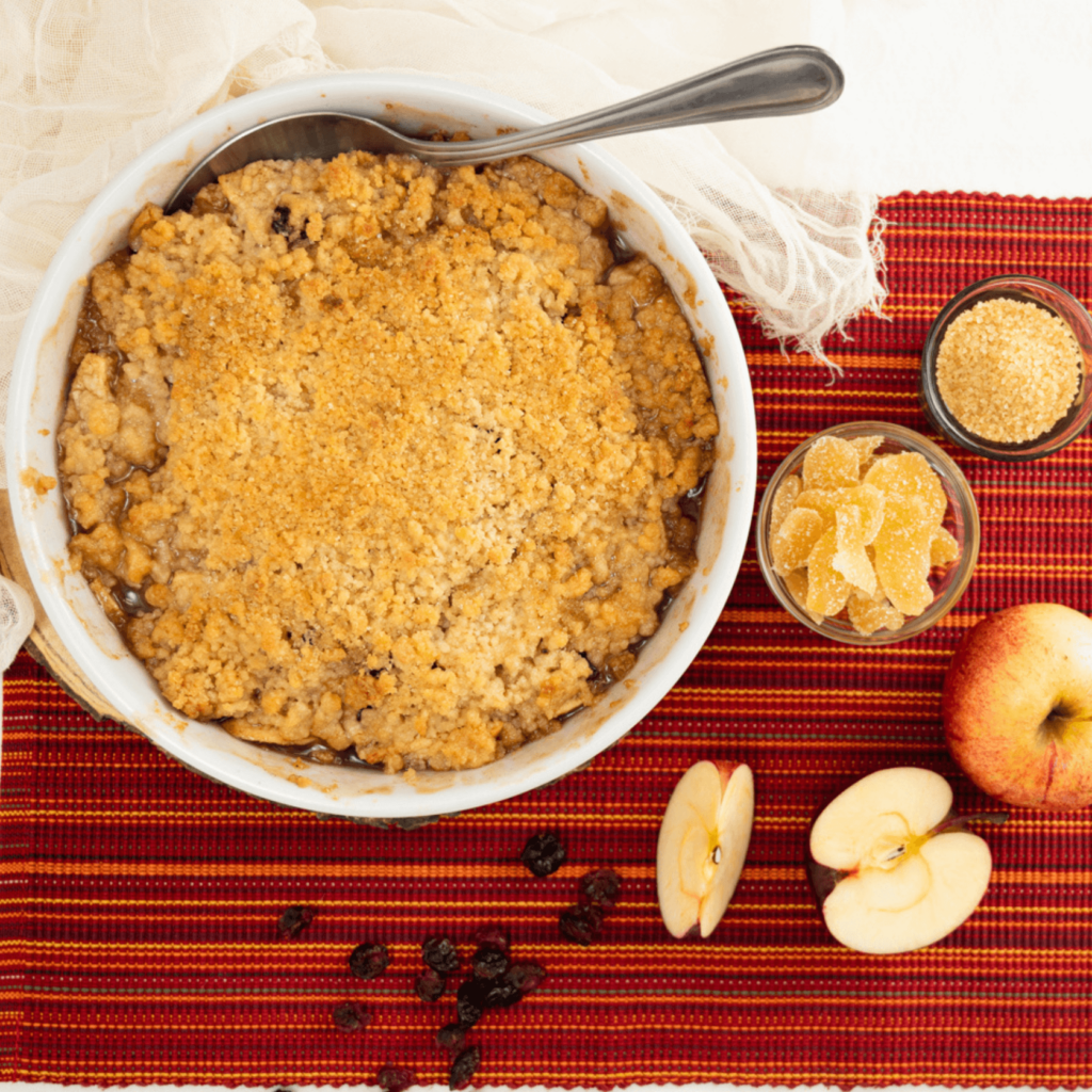 The image is shot from above and shows a round casserole dish with the apple crumble on a striped red placemant. Clockwise the upper right is a small bowl of brown sugar, a small bowl of crystallized ginger, a whole apple, a half apple, a segment of apple, and dried cranberries.