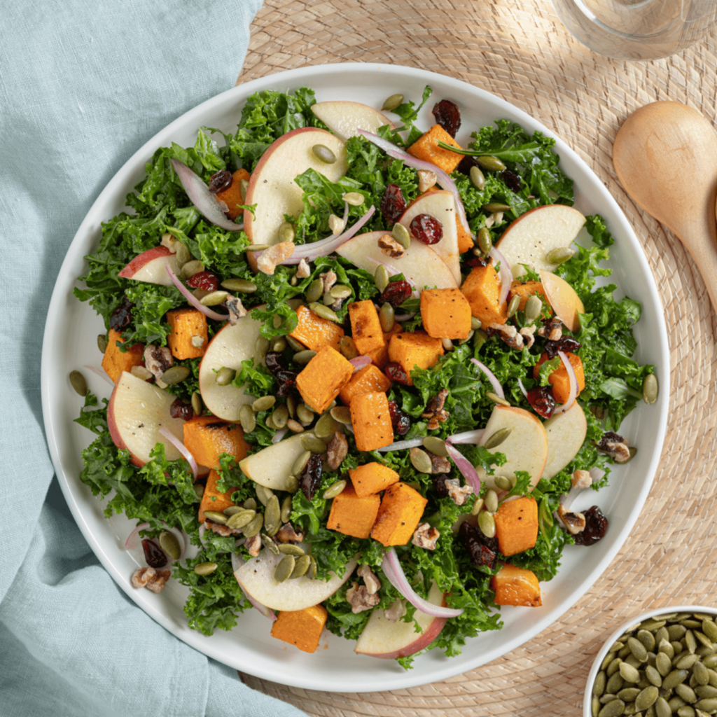 The image is shot from above and shows a large plate upon which is kale, thin slices of apple, cubes of butternut squash, thin slices of shallot, pumpkin seeds, and dried cranberries.