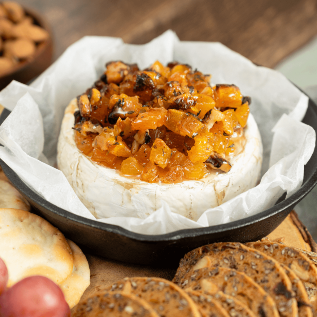 The image shows a small cast iron pan laid with parchment paper and a wheel of Brie. the Brie is topped with a compote of apricots, walnuts, and Ontario honey. The pan is surrounded by grapes and various crackers.