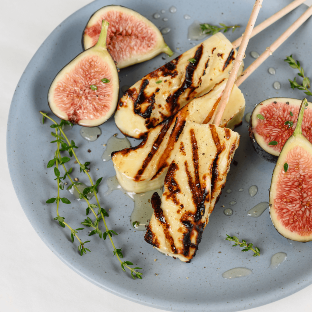 The image shows a blue plate with three wooden skewers of grilled halloumi cheese. The plate is garnished with fresh halved figs, drizzled honey, and sprigs of thyme.