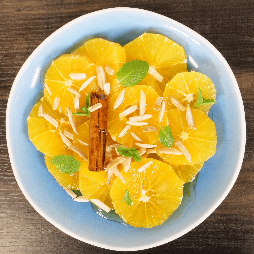 The image shows a top view of a blue plate with sliced and peeled wheels of oranges. The oranges are garnished with mint leaves, slivered almonds, and a cinnamon stick.