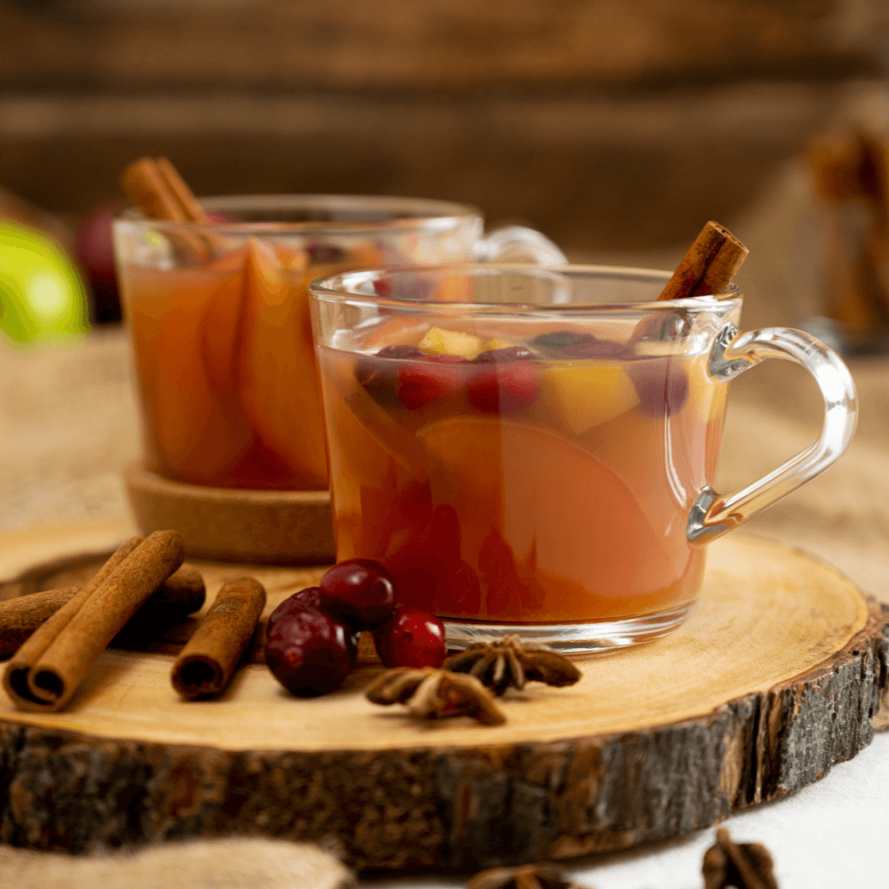 The image shows two clear glass mugs of mulled apple cider on a cross-section of a piece of wood. The mugs are garnished with whole cranberries and cinnamon sticks. In front of the mugs are cinnamon sticks, cranberries, and star anise.