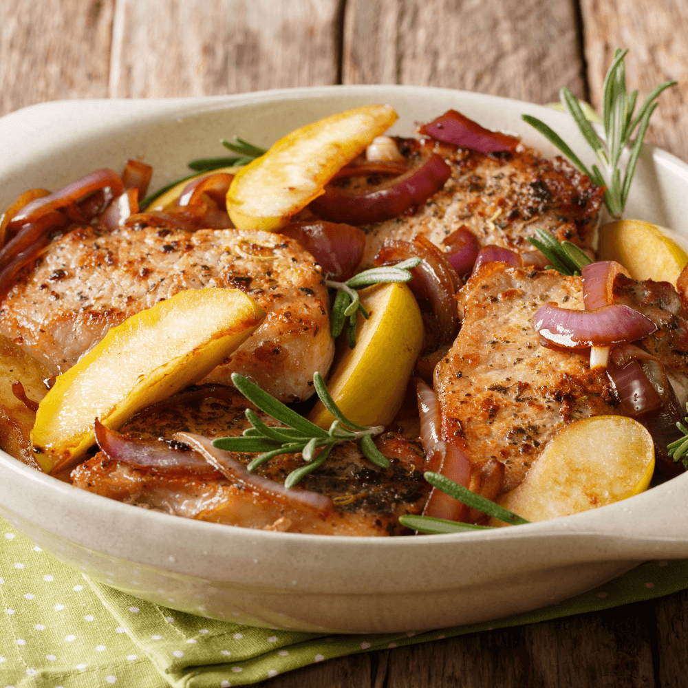 Close up of a white baking dish with baked pork chops, baked apples, cooked ed onions, and rosemary sprigs.