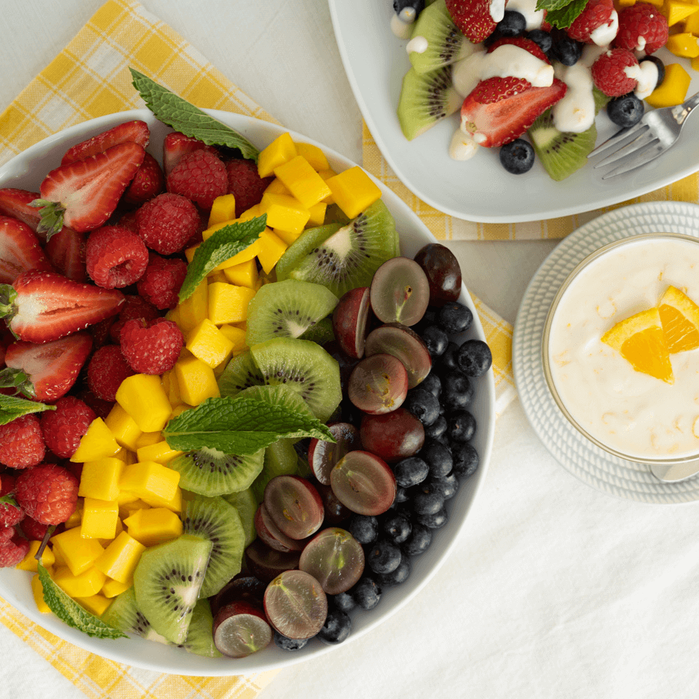 The images shows a top view of a fruit salad, arranged in rows of strawberries, raspberries, mango cubes, kiwi slices, halved red grapes, and blueberries. There is a small bowl of honey yogurt dipping sauce garnished with wedges of orange.