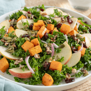 Plate with warm kale, apple, and butternut squash salad garnished with sliced red onions, pumpkin seeds, and dried cranberries.