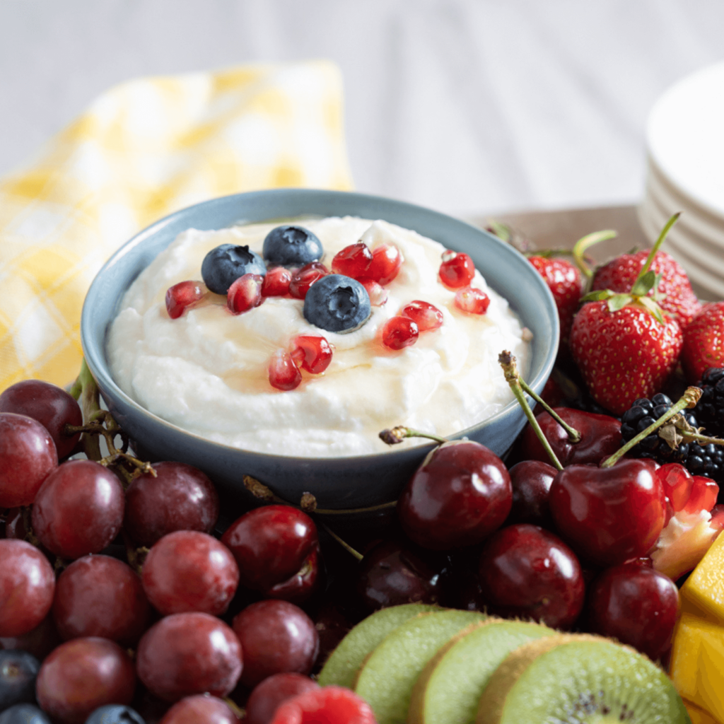 The image shows a small blue bowl filled with a whipped honey lemon ricotta dip. The dip is garnished with blueberries and pomegranate arils. The bowl is surrounded by grapes, strawberries, and sliced kiwi.