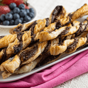 Oblong dish on pink napkin piled with chocolate puff pastry twists. A small bowl of strawberries and blueberries are in the background.