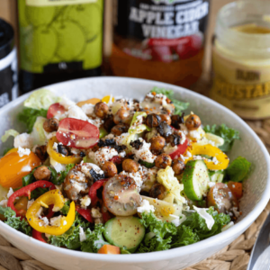 Angled view of a white bowl filled with salad in front of Farm Boy Fleur de Sel, Farm Boy Extra Virgin Olive Oil, Farm Boy Organic Apple Cider Vinegar, and Farm Boy Dijon Mustard