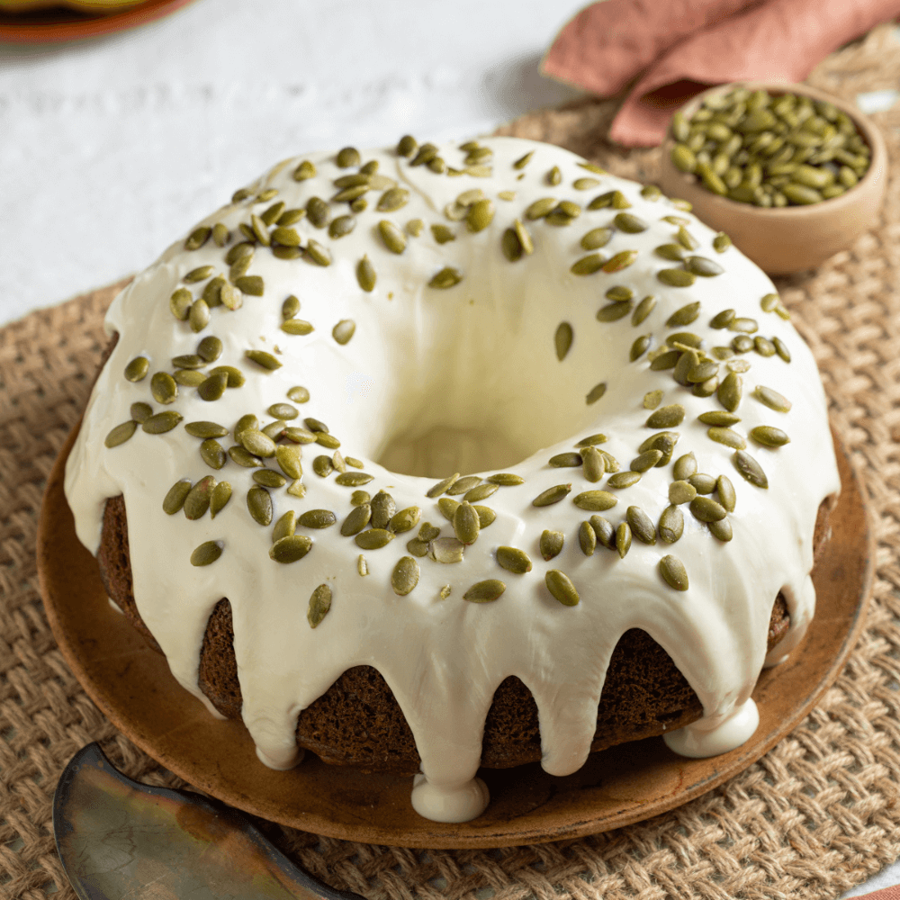 The image shows a Bundt cake on a plate covered in a cream cheese glaze and garnished with pumpkin seeds.