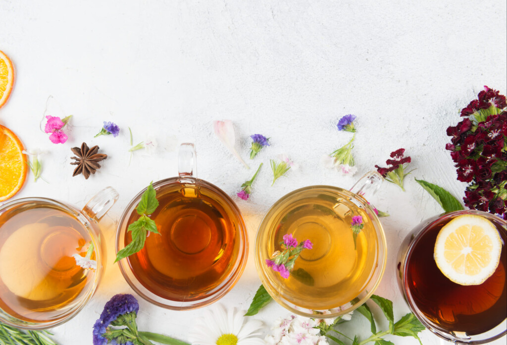 Directly above view of a four tea cups with different herbal teas. Anise, flowers and dried orange slices are spread around the tea cups on white background. One of the teas has a lemon slice.